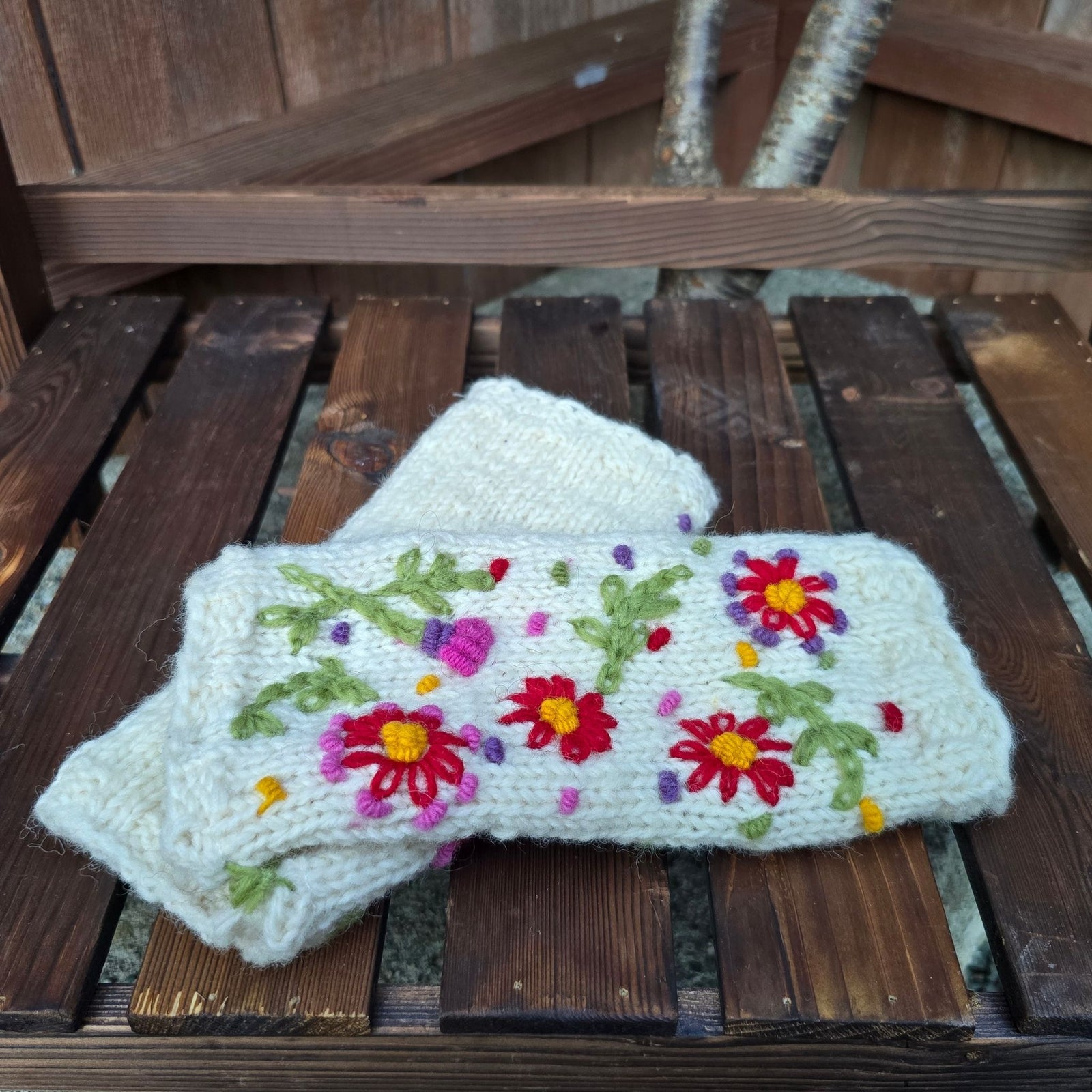Cream hand knitted wool handwarmers with red daisy floral embroidery handmade in Nepal laid on wooden surface